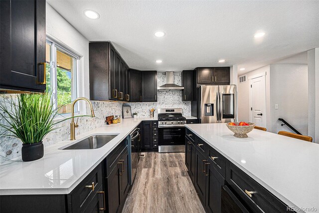 6032 Estes St, Arvada, CO 80004 - kitchen with appliances with stainless steel finishes, wall chimney range hood, dark cabinetry, light wood finished floors, and recessed lighting