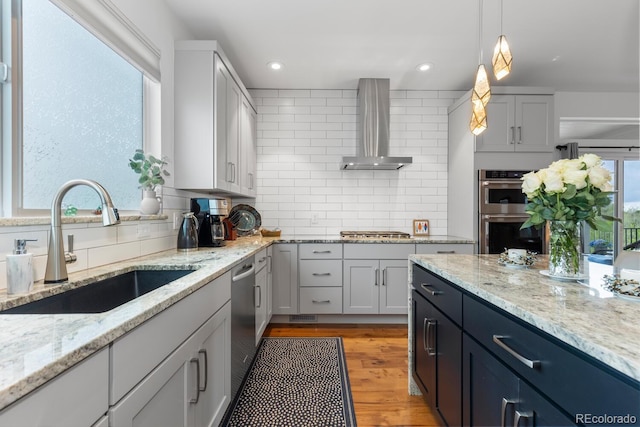 kitchen featuring wall chimney exhaust hood, stainless steel appliances, a sink, backsplash, and decorative light fixtures