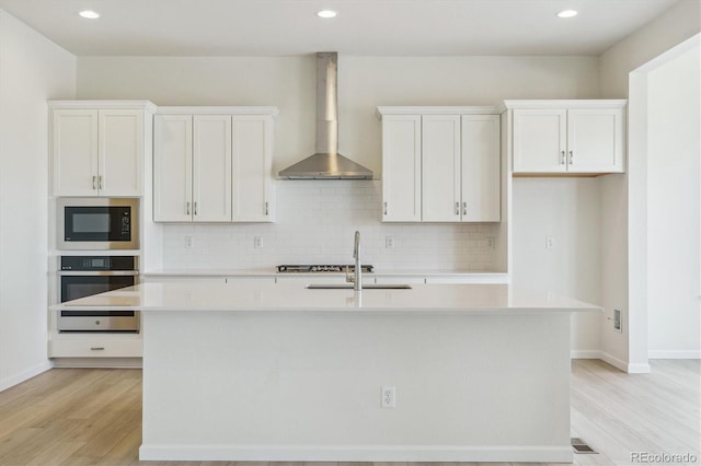 kitchen with stainless steel appliances, white cabinetry, wall chimney exhaust hood, and an island with sink