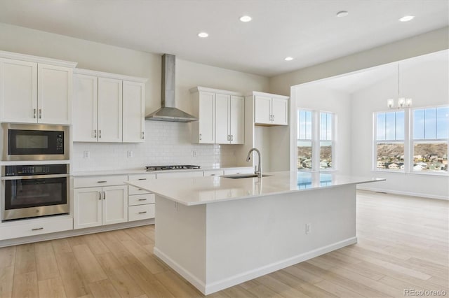 kitchen with wall chimney exhaust hood, stainless steel appliances, sink, white cabinetry, and an island with sink