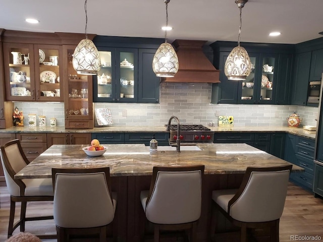 kitchen with a center island with sink, decorative light fixtures, light stone counters, and premium range hood