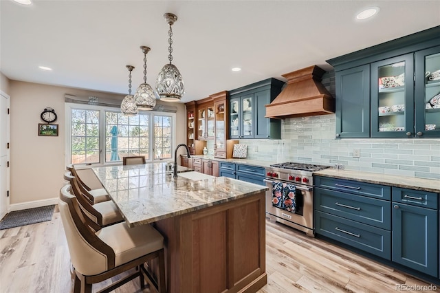 kitchen with custom exhaust hood, designer range, sink, light hardwood / wood-style flooring, and an island with sink