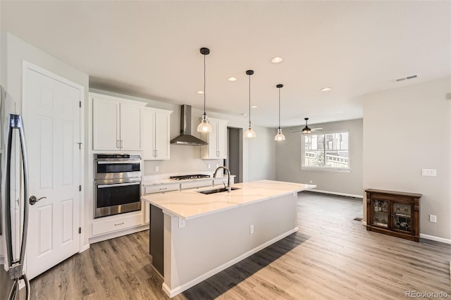 kitchen featuring a kitchen island with sink, white cabinets, wall chimney range hood, sink, and decorative light fixtures