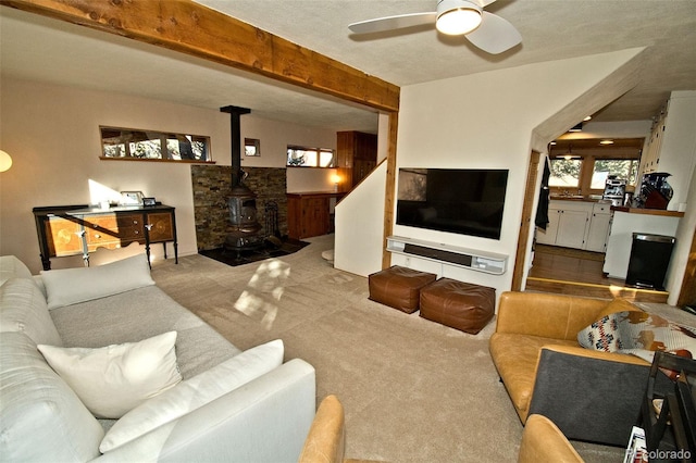 carpeted living room with a wood stove, ceiling fan, beamed ceiling, and a textured ceiling