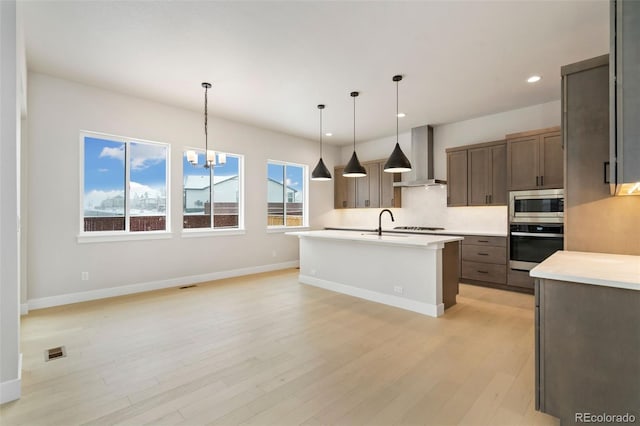 kitchen with wall chimney exhaust hood, stainless steel appliances, a kitchen island with sink, pendant lighting, and light hardwood / wood-style flooring