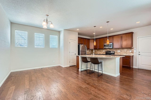 kitchen featuring appliances with stainless steel finishes, pendant lighting, backsplash, a kitchen island with sink, and dark wood-type flooring