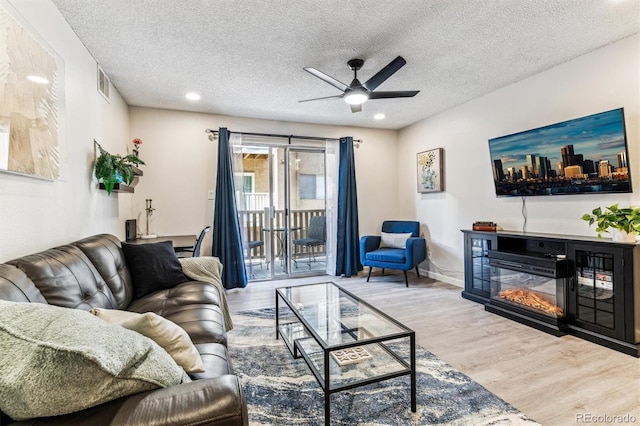 living area featuring visible vents, a ceiling fan, a glass covered fireplace, wood finished floors, and a textured ceiling