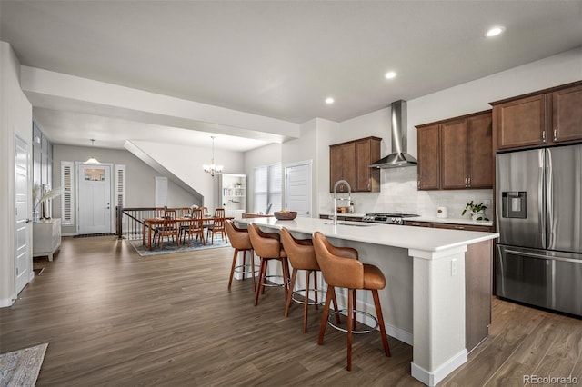 kitchen featuring wall chimney range hood, a kitchen bar, dark wood-style flooring, stainless steel fridge, and a kitchen island with sink