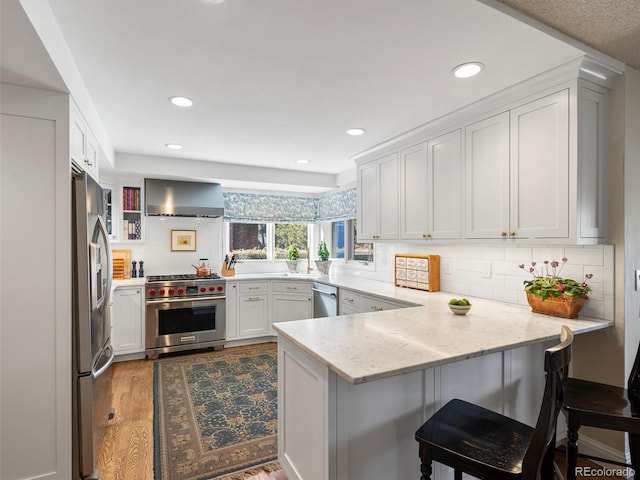 kitchen featuring wall chimney exhaust hood, appliances with stainless steel finishes, a kitchen breakfast bar, a peninsula, and white cabinetry