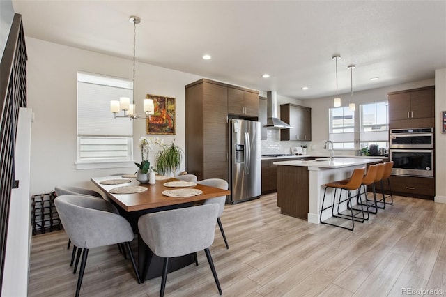 kitchen featuring wall chimney exhaust hood, stainless steel appliances, a kitchen breakfast bar, a center island with sink, and a notable chandelier
