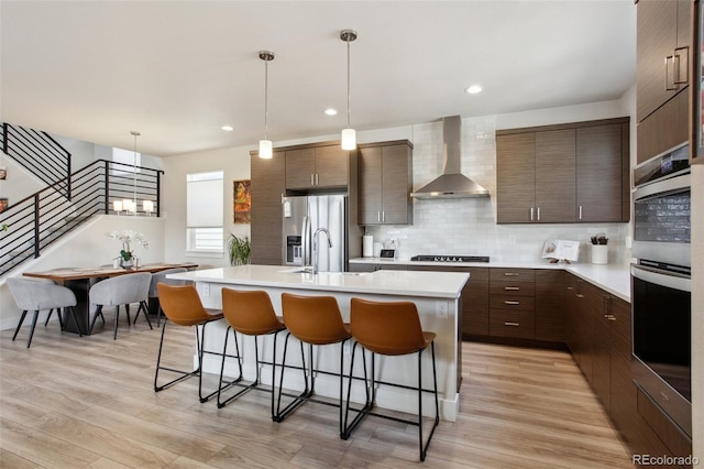kitchen with a kitchen island with sink, stainless steel appliances, light countertops, a kitchen bar, and wall chimney exhaust hood