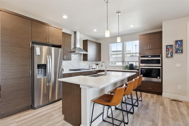 kitchen featuring a breakfast bar, a sink, light wood-type flooring, wall chimney exhaust hood, and appliances with stainless steel finishes