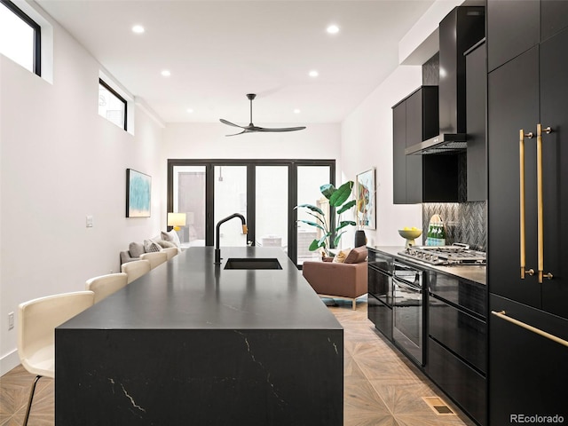 kitchen featuring backsplash, wall chimney range hood, sink, an island with sink, and stainless steel gas cooktop