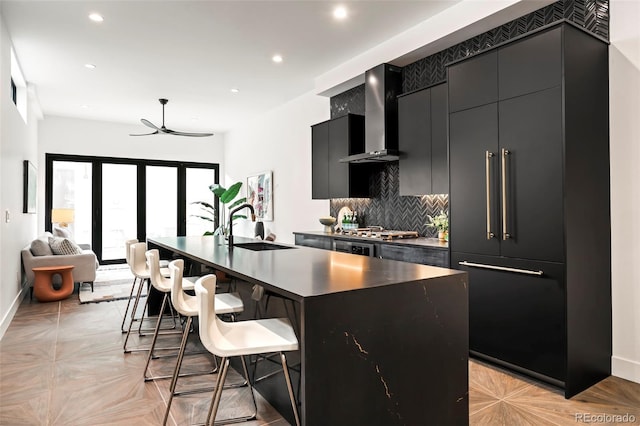 kitchen featuring a kitchen bar, tasteful backsplash, sink, wall chimney range hood, and an island with sink