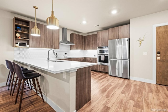 kitchen featuring wall chimney range hood, sink, appliances with stainless steel finishes, hanging light fixtures, and a kitchen breakfast bar