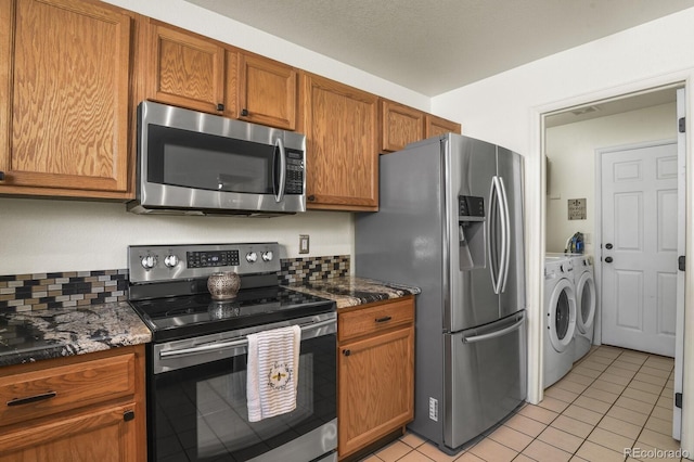 kitchen with brown cabinets, dark stone countertops, washing machine and dryer, light tile patterned flooring, and appliances with stainless steel finishes