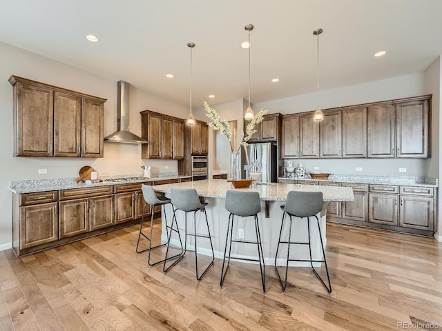 kitchen featuring wall chimney exhaust hood, light stone counters, decorative light fixtures, a center island with sink, and stainless steel appliances