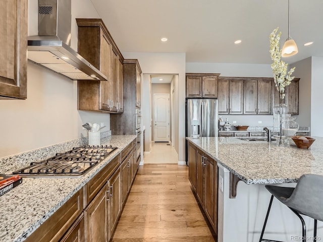 kitchen featuring wall chimney range hood, stainless steel appliances, a kitchen breakfast bar, and light stone countertops