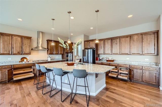 kitchen with wall chimney exhaust hood, hanging light fixtures, stainless steel appliances, light stone countertops, and a kitchen island with sink