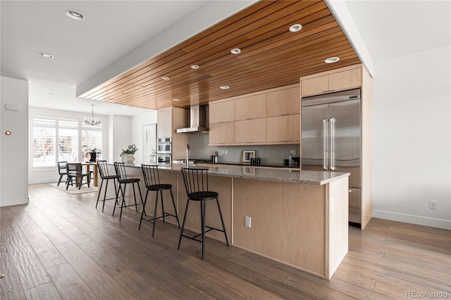 kitchen with stone countertops, appliances with stainless steel finishes, wood ceiling, a kitchen island with sink, and wall chimney range hood