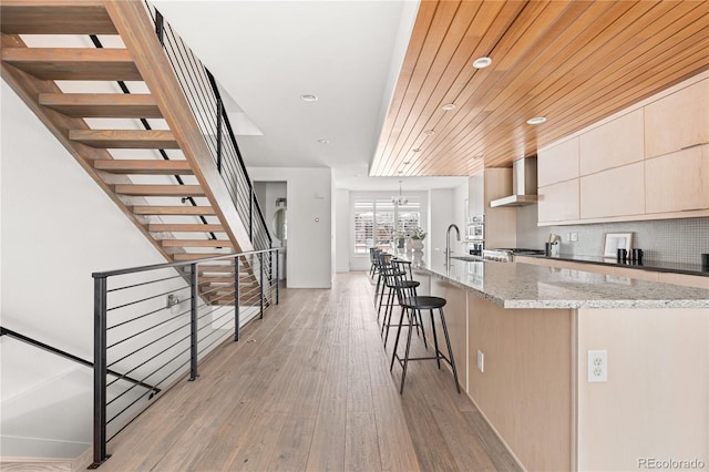 kitchen featuring decorative light fixtures, light stone counters, wall chimney range hood, light hardwood / wood-style floors, and decorative backsplash