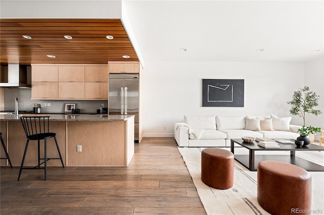 kitchen with stainless steel built in refrigerator, wall chimney exhaust hood, light wood-type flooring, dark stone counters, and wooden ceiling