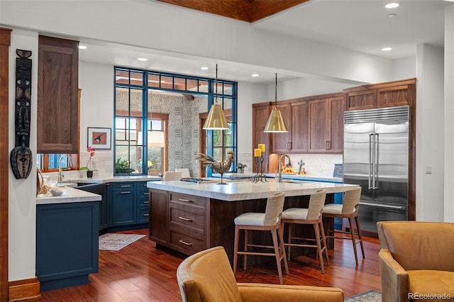 kitchen featuring decorative light fixtures, stainless steel built in refrigerator, backsplash, dark hardwood / wood-style flooring, and a kitchen island with sink