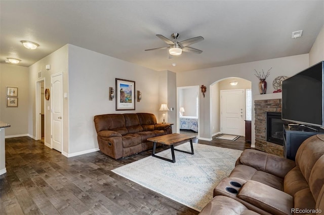 living room with a stone fireplace, dark wood-type flooring, and ceiling fan