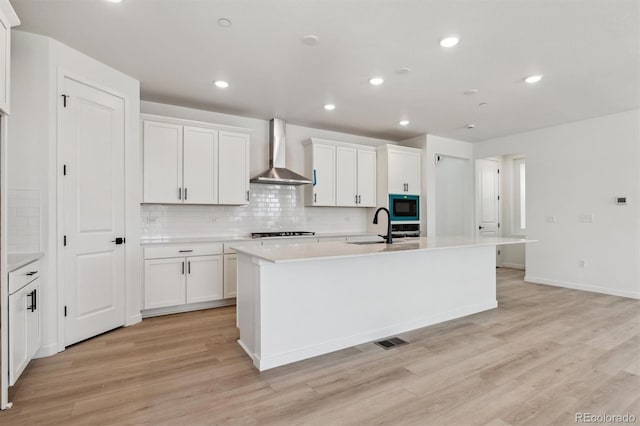 kitchen with light wood finished floors, a sink, wall chimney exhaust hood, a kitchen island with sink, and tasteful backsplash