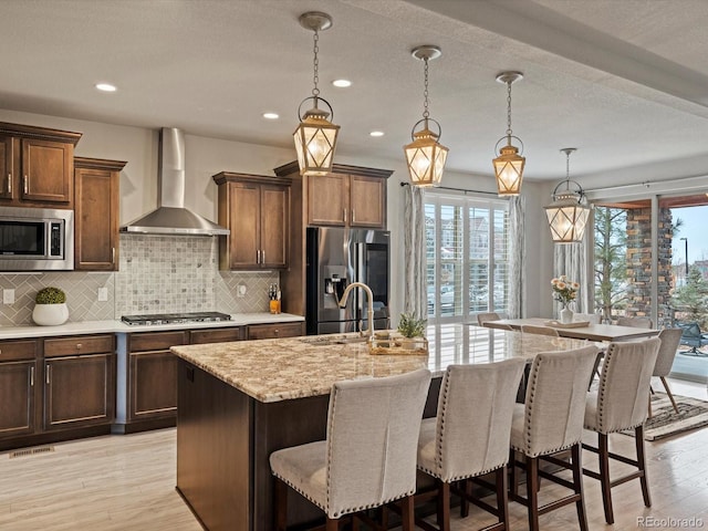 kitchen featuring appliances with stainless steel finishes, decorative light fixtures, a kitchen island with sink, wall chimney range hood, and a sink