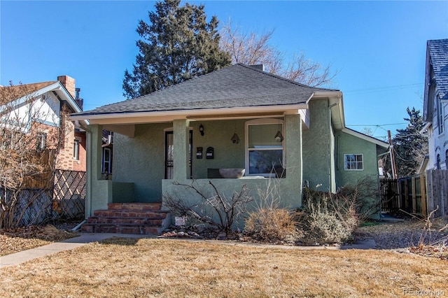 bungalow featuring roof with shingles, covered porch, fence, a front lawn, and stucco siding