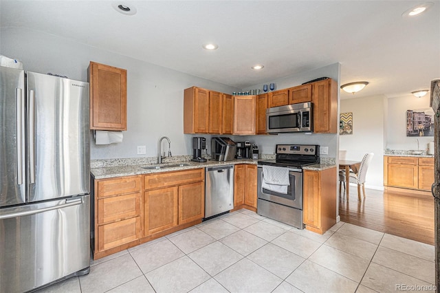 kitchen featuring light tile patterned floors, brown cabinetry, appliances with stainless steel finishes, light stone countertops, and a sink