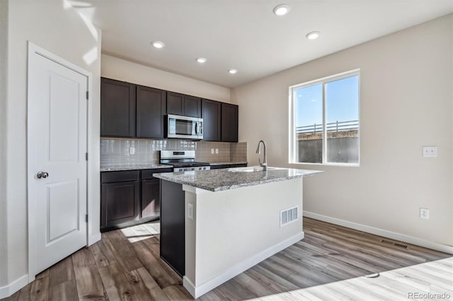 kitchen featuring light stone countertops, dark brown cabinets, stainless steel appliances, sink, and a center island with sink