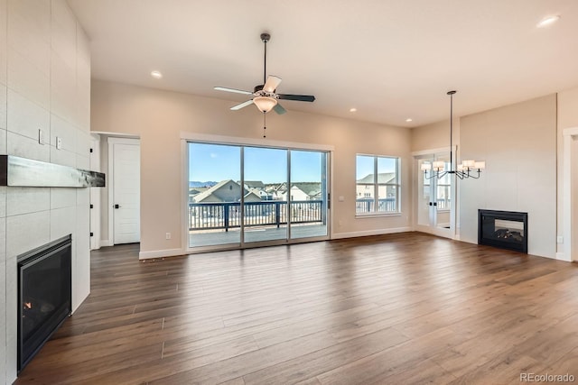 unfurnished living room featuring dark wood-style flooring, recessed lighting, a tiled fireplace, baseboards, and ceiling fan with notable chandelier