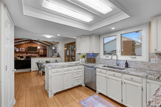 kitchen with a tray ceiling, a peninsula, a sink, light wood-style floors, and stainless steel dishwasher