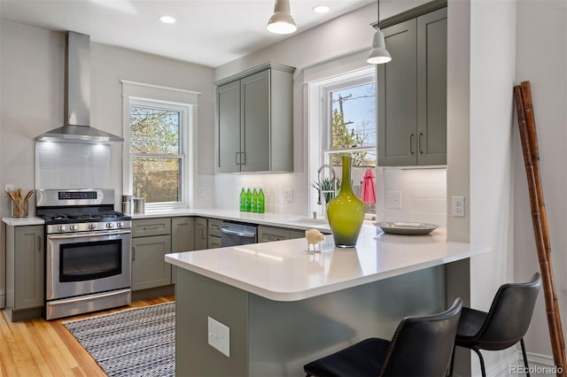 kitchen with kitchen peninsula, wall chimney exhaust hood, hanging light fixtures, decorative backsplash, and appliances with stainless steel finishes