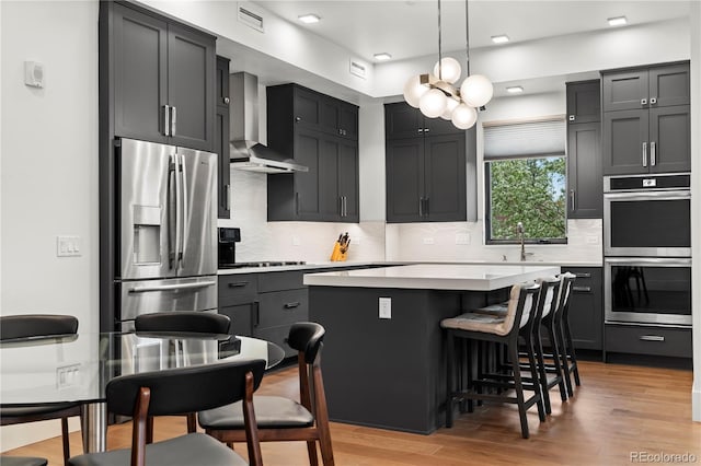 kitchen with appliances with stainless steel finishes, hanging light fixtures, a center island, wall chimney range hood, and light wood-type flooring