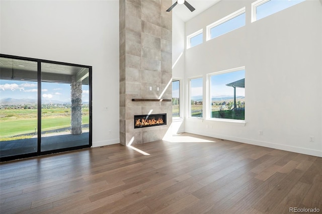 unfurnished living room featuring a tiled fireplace, wood finished floors, a high ceiling, baseboards, and ceiling fan