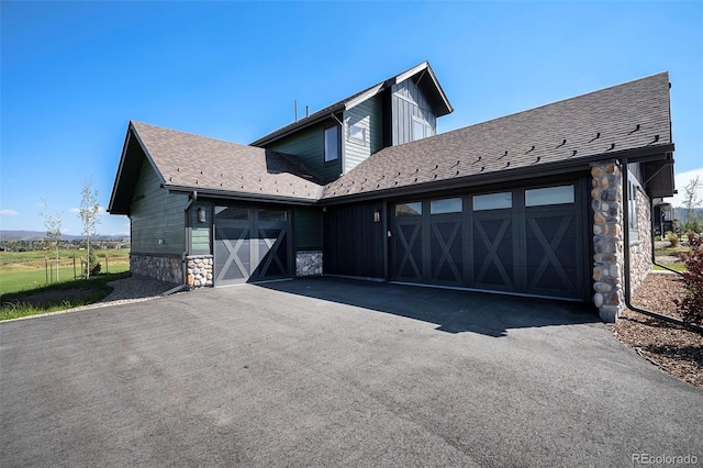 exterior space with aphalt driveway, stone siding, a garage, and roof with shingles