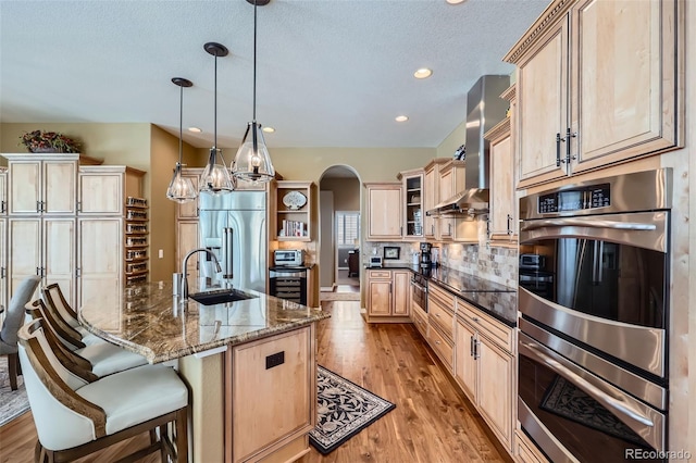 kitchen with decorative light fixtures, sink, dark stone countertops, stainless steel appliances, and wall chimney range hood