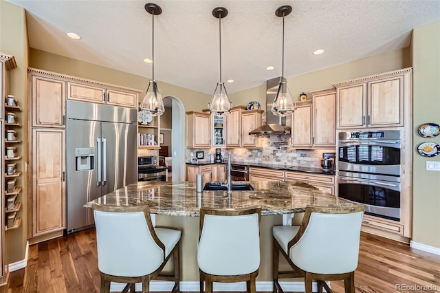 kitchen featuring wall chimney exhaust hood, pendant lighting, an island with sink, hardwood / wood-style flooring, and stainless steel appliances