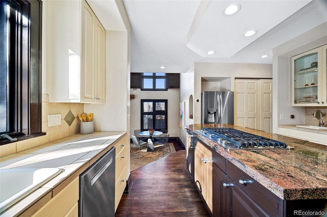 kitchen with cream cabinetry, dark brown cabinets, appliances with stainless steel finishes, and glass insert cabinets