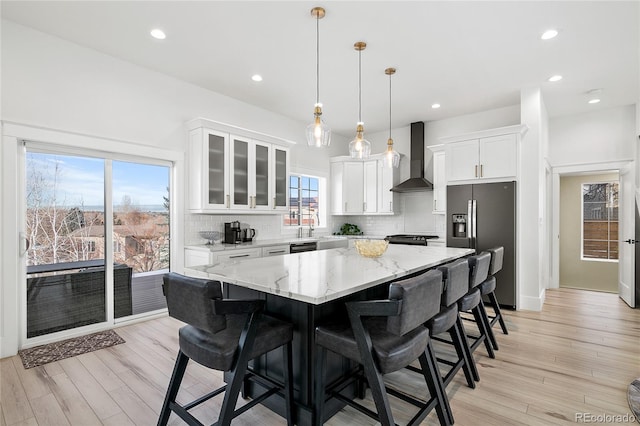 kitchen with pendant lighting, wall chimney range hood, a kitchen island, white cabinetry, and appliances with stainless steel finishes