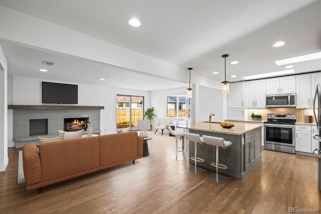 kitchen with a center island with sink, hanging light fixtures, a brick fireplace, tasteful backsplash, and stainless steel appliances