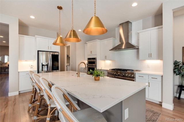 kitchen featuring white cabinets, wall chimney exhaust hood, decorative light fixtures, stainless steel appliances, and a kitchen island with sink