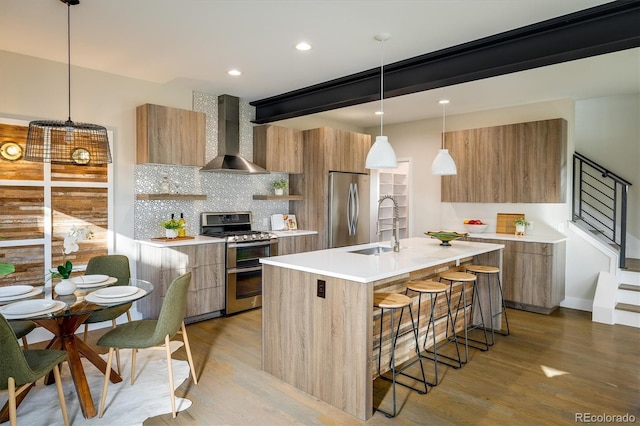 kitchen with pendant lighting, wall chimney exhaust hood, an island with sink, appliances with stainless steel finishes, and tasteful backsplash