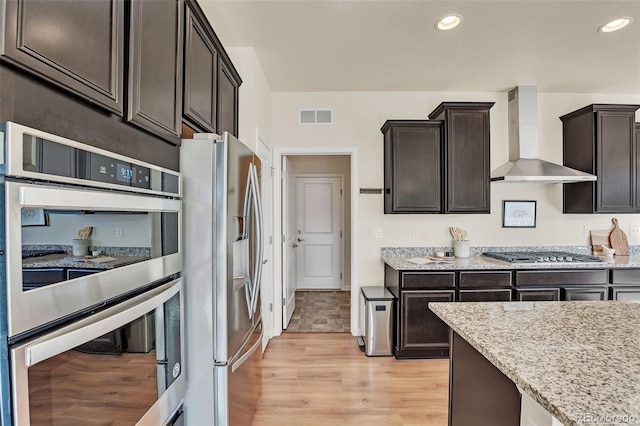 kitchen with wall chimney range hood, light stone countertops, stainless steel appliances, and visible vents