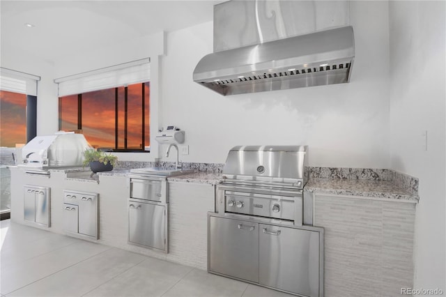 kitchen with light tile patterned flooring, wall chimney range hood, light stone counters, and a sink