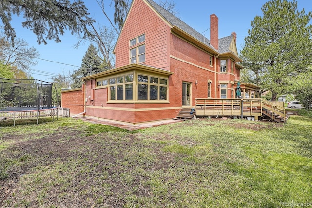rear view of property with a trampoline, a deck, a chimney, brick siding, and a lawn