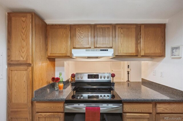 kitchen with under cabinet range hood, stainless steel electric stove, brown cabinetry, and dark stone countertops
